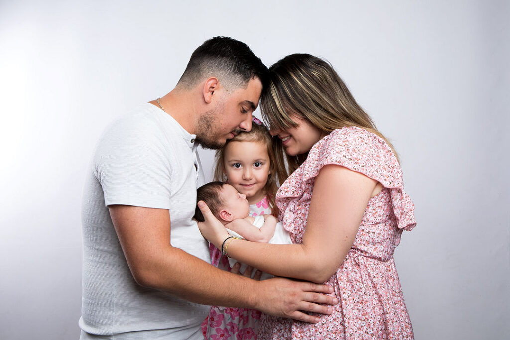 Un portrait de famille plein d'amour, où une mère et un père enlacent leurs deux enfants. Le père, en chemise blanche, tient un nouveau-né, tandis que sa sœur aînée sourit. La mère, vêtue d'une robe rose à fleurs, serre la famille dans ses bras, lui transmettant amour et chaleur.