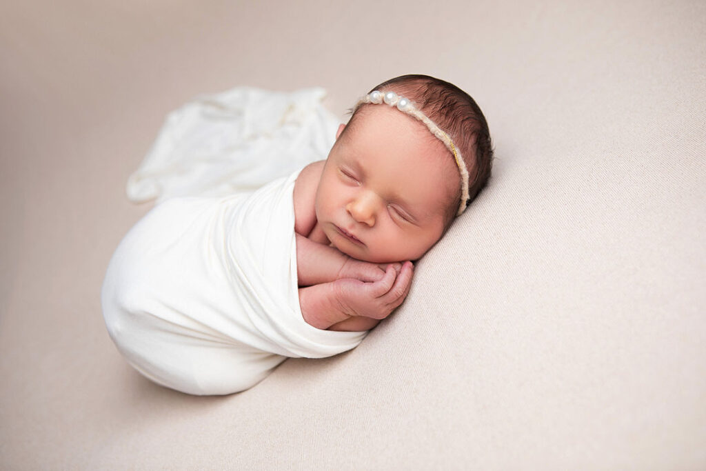 Un nouveau-né paisiblement endormi, emmailloté dans un doux tissu blanc, repose sur une couverture beige. Il porte un délicat serre-tête orné de perles, une pose classique pour la photographie de nouveau-nés.