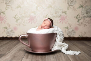 Un nouveau-né endormi dans une tasse de thé rose doré, enveloppé dans une douce couverture blanche. Idée de séance photo pour bébé avec un fond floral. Accessoire idéal pour la photographie de nouveau-né.