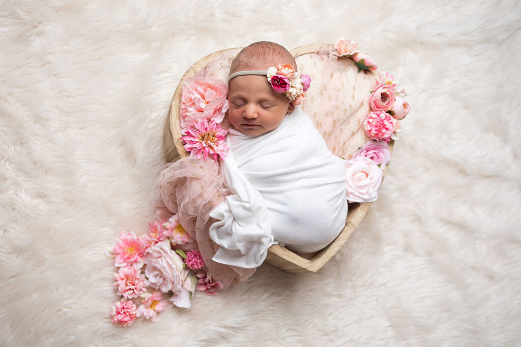 Un nouveau-né dort paisiblement, emmailloté de blanc, blotti dans un bol en bois en forme de cœur, entouré de fleurs roses et pêche. Un délicat bandeau fleuri orne sa tête, posée sur un tapis blanc moelleux, idéal pour une séance photo de nouveau-né.