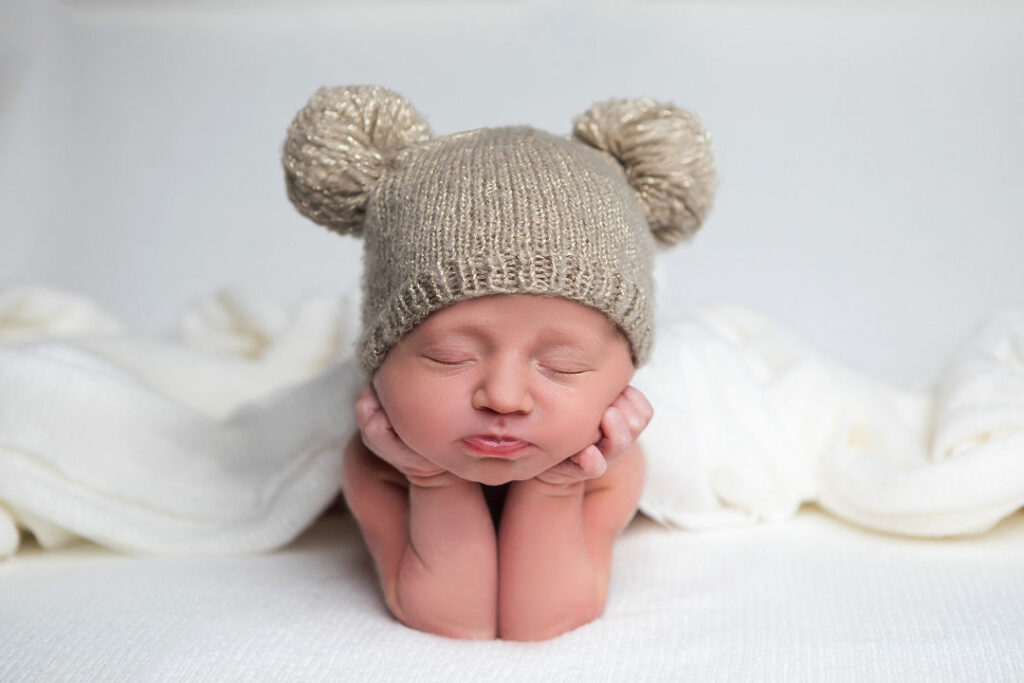 Un nouveau-né serein, coiffé d'un bonnet beige tricoté aux grandes oreilles rondes, repose paisiblement, les mains sous le menton. Des couvertures blanches douces et un fond texturé créent un décor douillet pour une séance photo de nouveau-né.