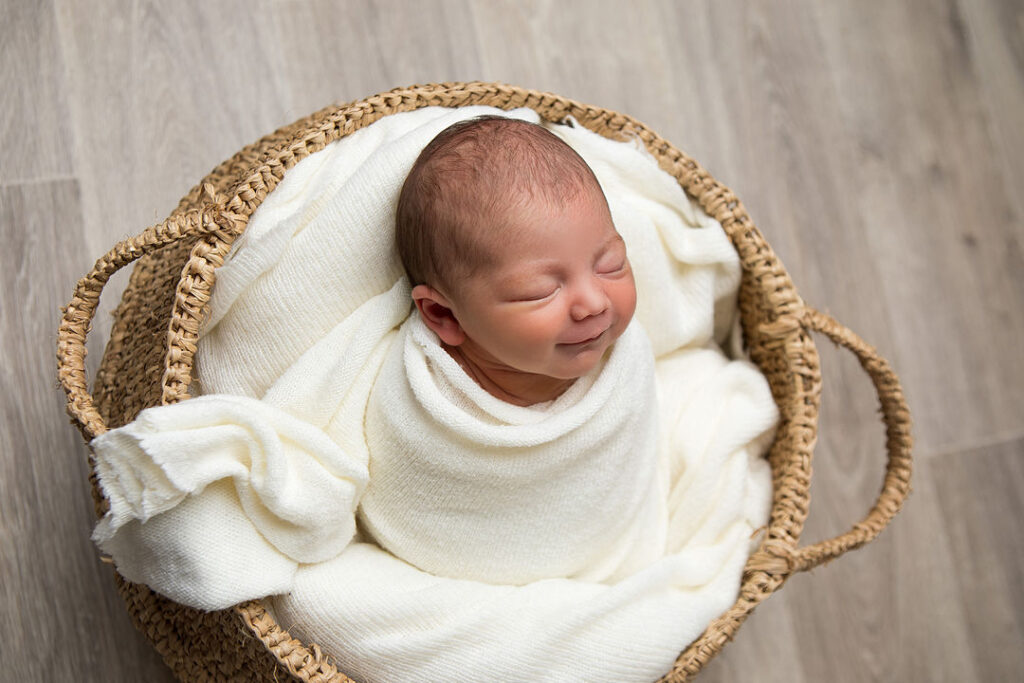 Un nouveau-né souriant, emmitouflé dans une douce couverture blanche, blotti dans un panier tressé. Une photo de bébé sereine et paisible, parfaite pour une séance photo de nouveau-né.