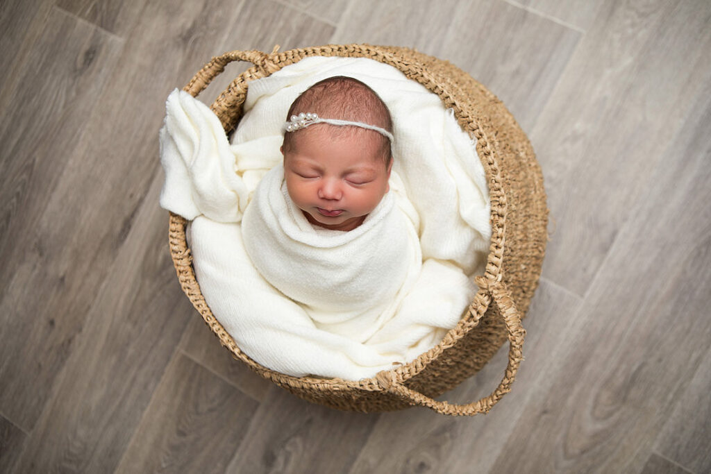 Un nouveau-né paisiblement emmitouflé dans une couverture blanche, blotti dans un panier tressé, crée une scène sereine pour la photographie de nouveau-né. La lumière douce et les tons naturels mettent en valeur les traits délicats du bébé, parfait pour les photos de bébé et une séance photo de nouveau-né.