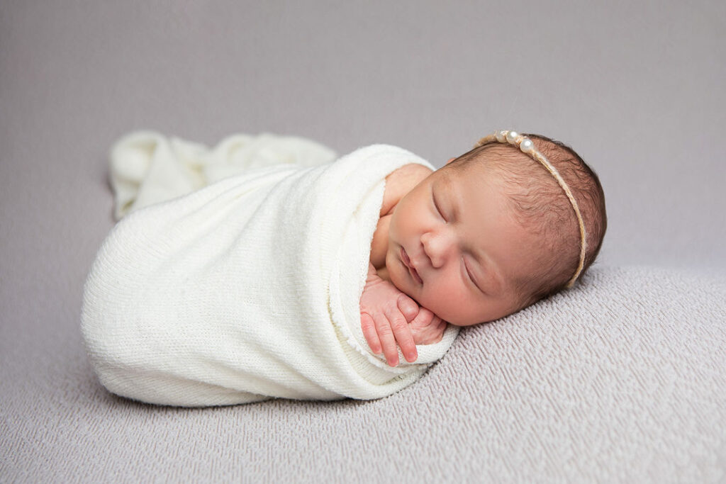 Un nouveau-né endormi, emmitouflé dans une couverture blanche, repose sur une surface grise et douce. Ce bébé, photographié lors d'une séance photo de nouveau-né, porte un bandeau de perles, créant une photo sereine et paisible.