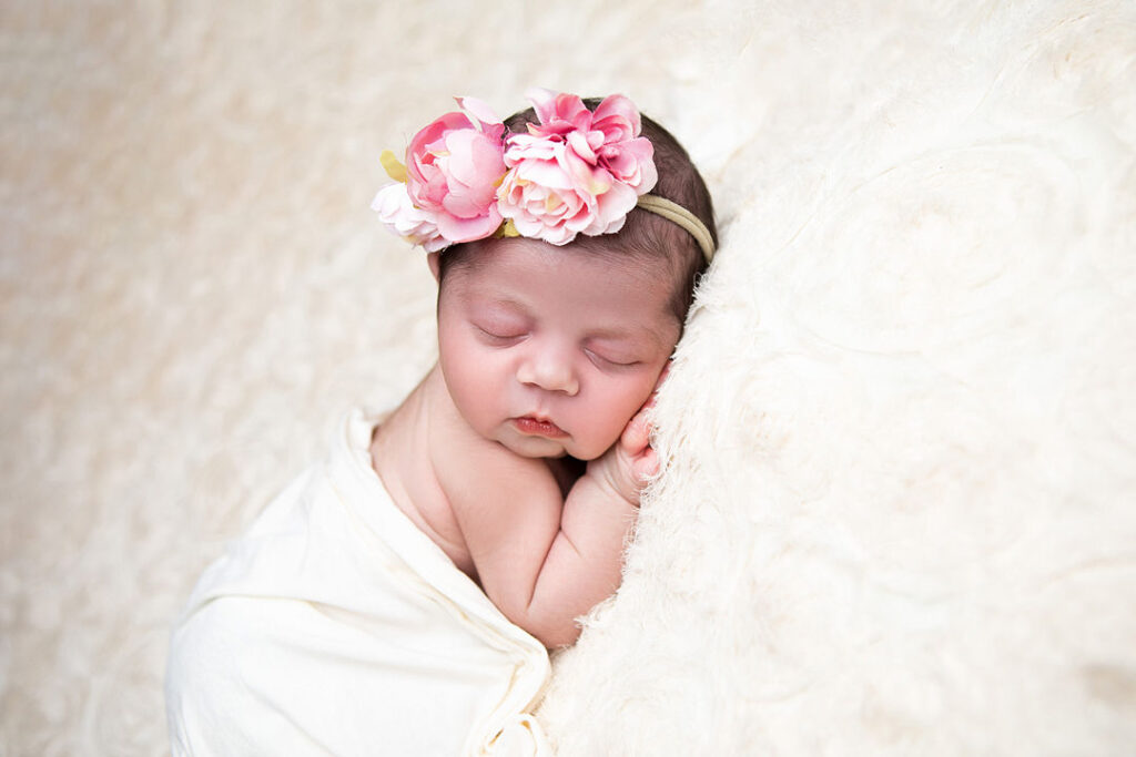 Un nouveau-né paisiblement endormi, enveloppé dans une couverture couleur crème, porte un bandeau à fleurs roses. Un fond doux et moelleux, idéal pour la photographie de nouveau-né.