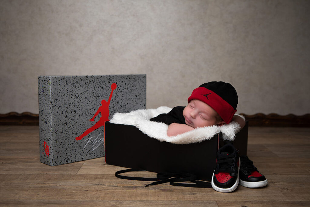 Un nouveau-né dort paisiblement dans une boîte à chaussures noire, coiffé d'un bonnet noir et rouge, avec de minuscules baskets Jordan et une boîte à chaussures assortie à ses côtés. Un décor parfait pour une séance photo de nouveau-né.