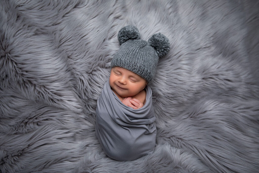 Nouveau-né souriant enveloppé dans une couverture grise, portant un bonnet gris tricoté avec des oreilles à pompon, allongé paisiblement sur un tapis en fourrure grise douce, parfait pour la photographie de nouveau-né et les photos de bébé.