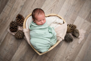 Un nouveau-né paisiblement endormi, emmailloté dans une douce couverture vert menthe, blotti dans un bol en bois en forme de cœur. Entouré de pommes de pin et de couvertures moelleuses, il est parfait pour une séance photo de nouveau-né.