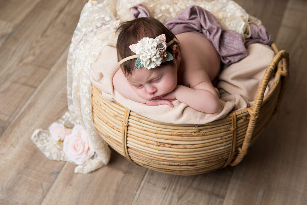Un nouveau-né dort paisiblement dans un panier tressé, coiffé d'un bandeau fleuri. Des couvertures douces et de la dentelle l'entourent, parfait pour une séance photo de nouveau-né. Photographie de bébé.
