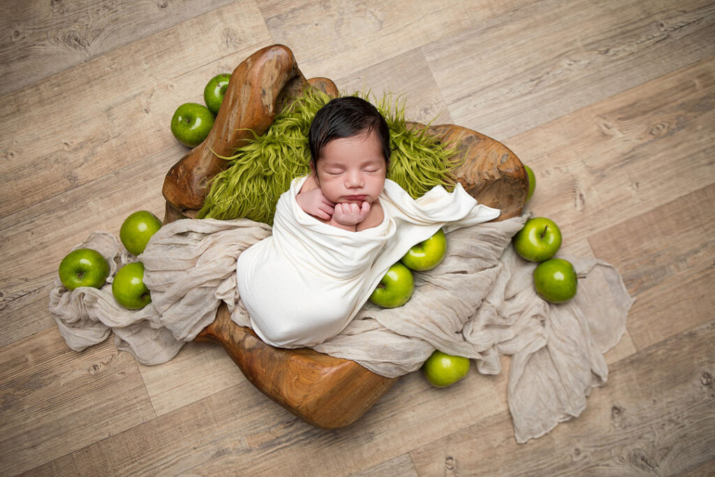 Nouveau-né emmailloté de blanc, blotti dans un bol en bois avec des pommes vertes et une couverture douce. Un décor de photographie de nouveau-né professionnel illustre une séance photo de bébé.