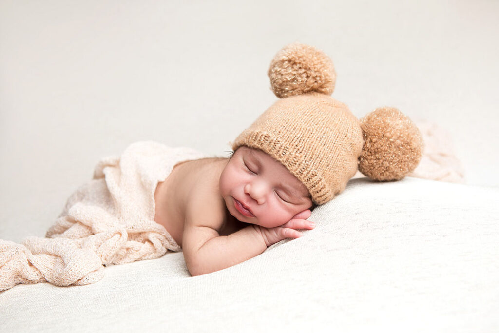 Un nouveau-né paisiblement endormi, coiffé d'un bonnet à pompons et enveloppé dans une douce couverture couleur crème. Il est photographié dans une pose classique, reposant sur une surface texturée.