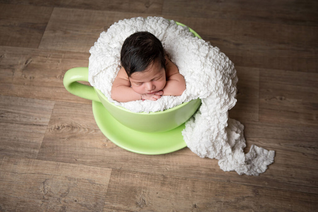 Un nouveau-né serein dort profondément, blotti dans une grande tasse de thé verte sur un parquet, enveloppé dans une douce couverture blanche. Cette photo de nouveau-né créative capture une séance photo paisible.
