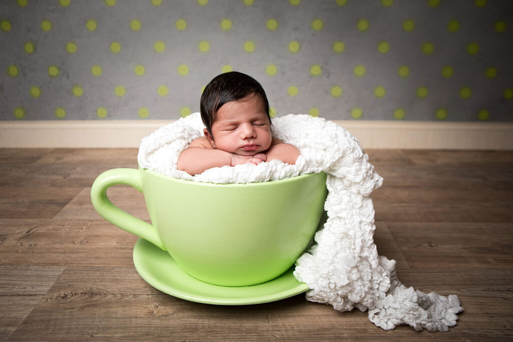 Un nouveau-né dort paisiblement dans une tasse de thé verte géante, enveloppé dans une douce couverture blanche. Adorable idée de séance photo pour nouveau-né.