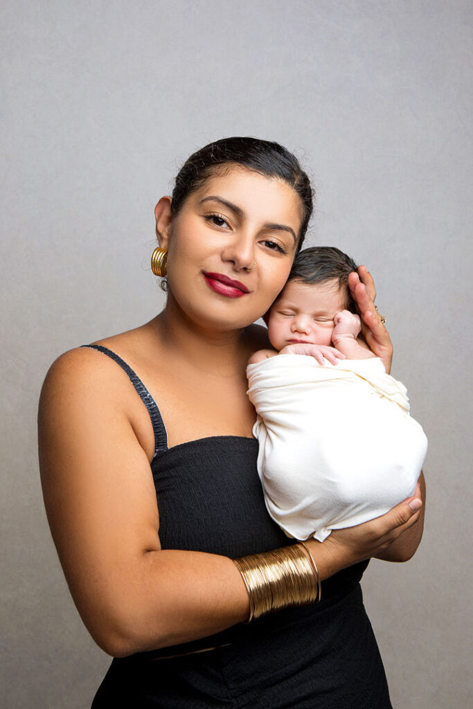 Une mère souriante berce tendrement un nouveau-né emmailloté dans ses bras. Elle porte une robe noire et des bijoux en or, et ses cheveux noirs sont tirés en arrière. Le bébé dort paisiblement, un moment parfait pour une séance photo de nouveau-né.
