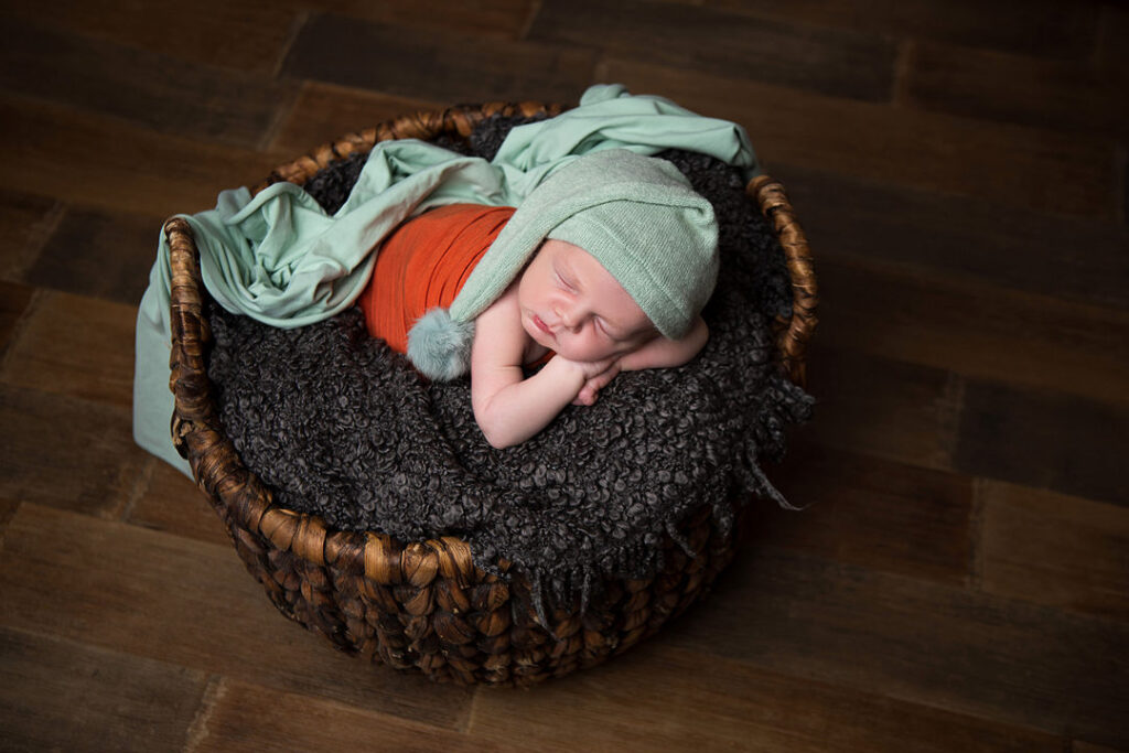 Un nouveau-né paisiblement endormi, emmailloté d'orange, coiffé d'un bonnet de lutin vert menthe à pompon, blotti dans un panier tressé sur un parquet. Cette séance photo de nouveau-né illustre une pose classique pour une séance photo de bébé.