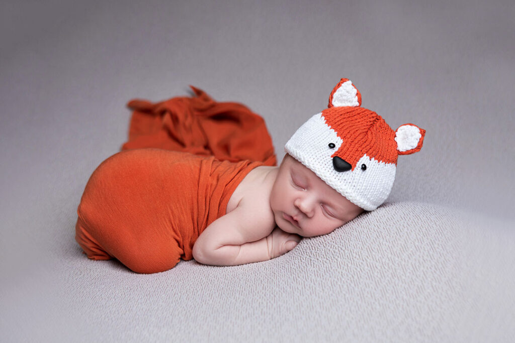 Un nouveau-né paisiblement endormi, emmailloté dans un tissu orange et coiffé d'un bonnet renard tricoté, repose sur une douce couverture aux tons neutres. Cette séance photo de nouveau-né sur le thème du renard est adorable.