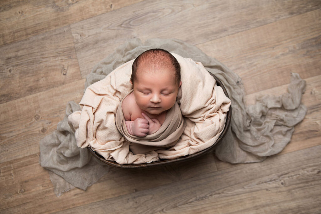 Nouveau-né paisiblement emmailloté dans une couverture beige, blotti dans un bol rustique, entouré de couvertures douces : parfait pour une séance photo de nouveau-né. Fond bois.