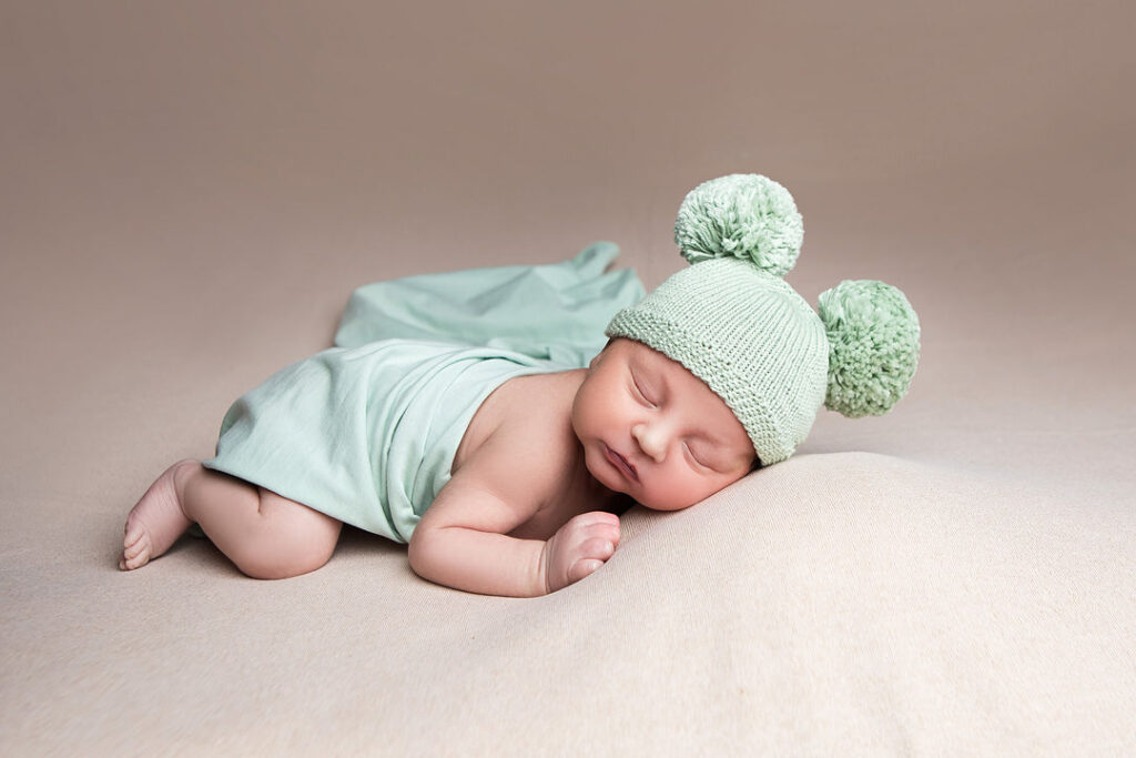 Un nouveau-né endormi, coiffé d'un bonnet vert menthe à pompons, enveloppé dans une couverture assortie, lors d'une séance photo de nouveau-né. Une lumière douce met en valeur les traits délicats de son visage, créant ainsi de magnifiques photos de bébé.
