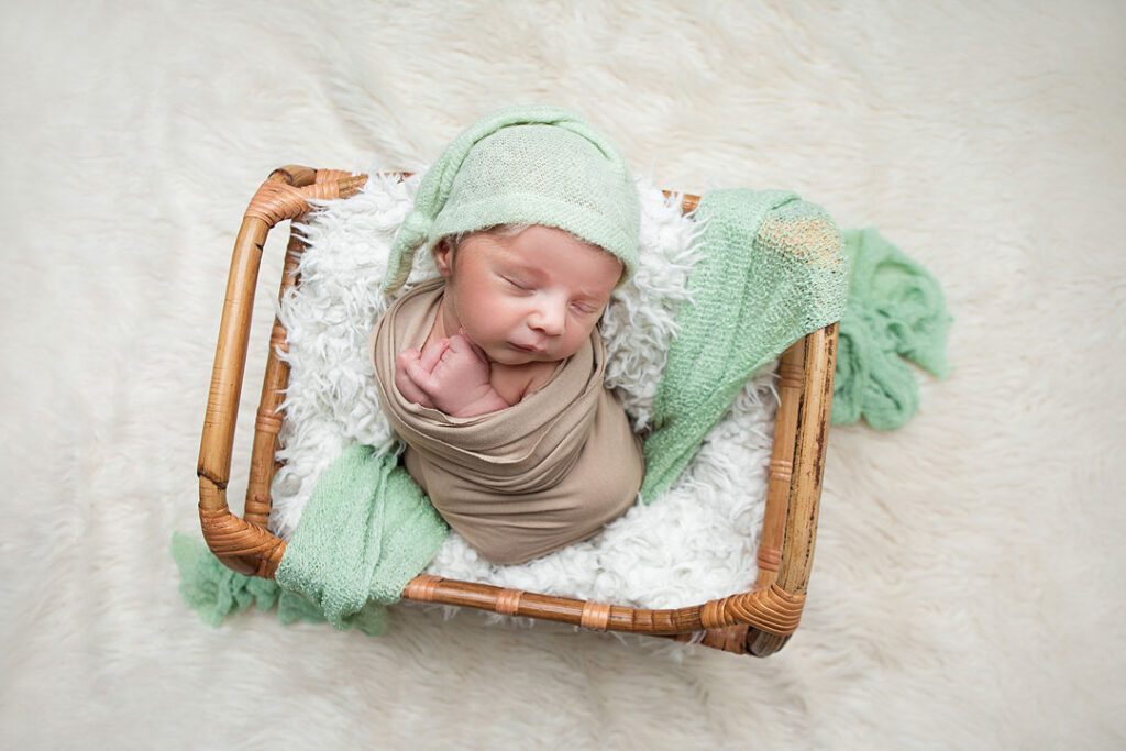 Nouveau-né endormi, emmailloté dans une couverture marron, coiffé d'un bonnet vert, blotti dans un panier tressé. Une douce couverture verte et une fourrure blanche moelleuse créent un cadre apaisant pour la photographie de nouveau-nés.