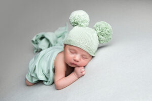 Un nouveau-né paisiblement endormi, enveloppé dans une douce couverture vert menthe, porte un bonnet assorti orné de pompons moelleux. Cette adorable séance photo capture la sérénité de la photographie de nouveau-né.