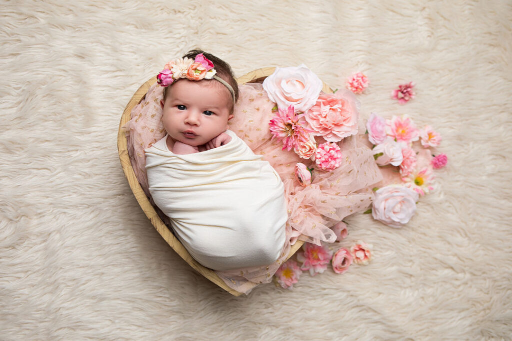 Un nouveau-né emmailloté avec un bandeau fleuri repose dans un bol en bois en forme de cœur, entouré de fleurs roses et d'une couverture blanche douce et moelleuse, parfaite pour la photographie de nouveau-né.