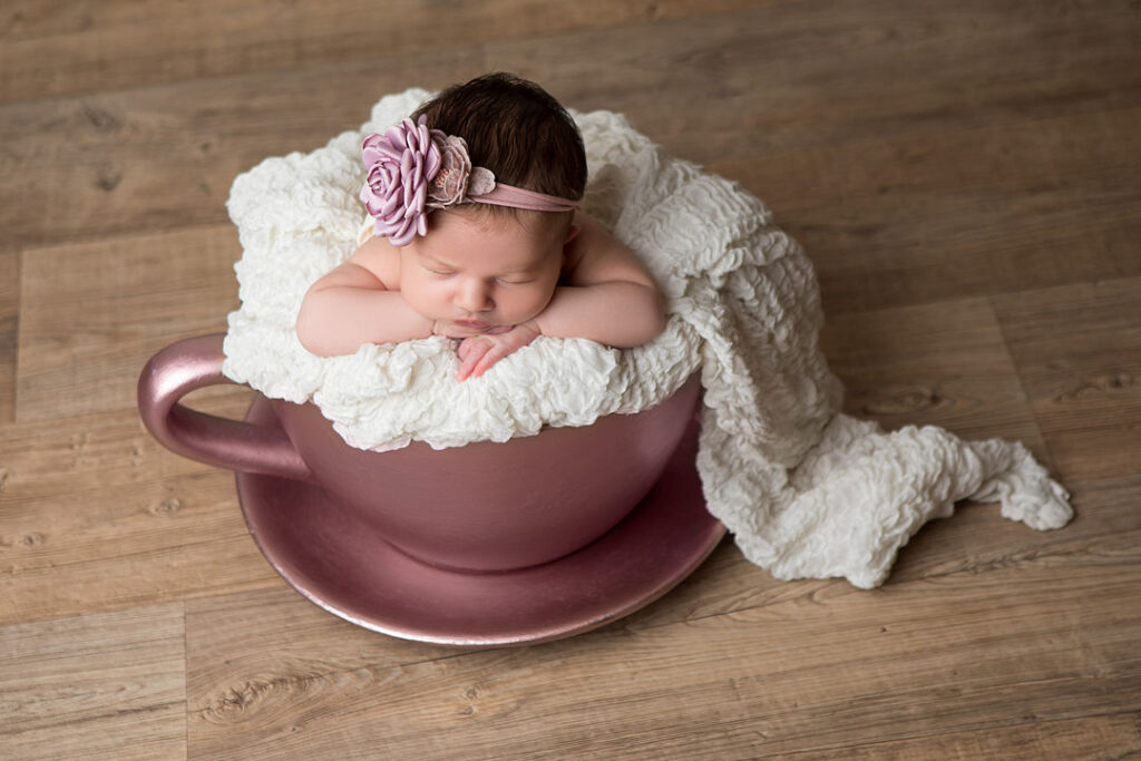 Une petite fille nouveau-née dort dans une grande tasse de thé rose, blottie dans une couverture blanche moelleuse. Elle porte un bandeau fleuri, une pose classique pour la photographie de nouveau-nés.