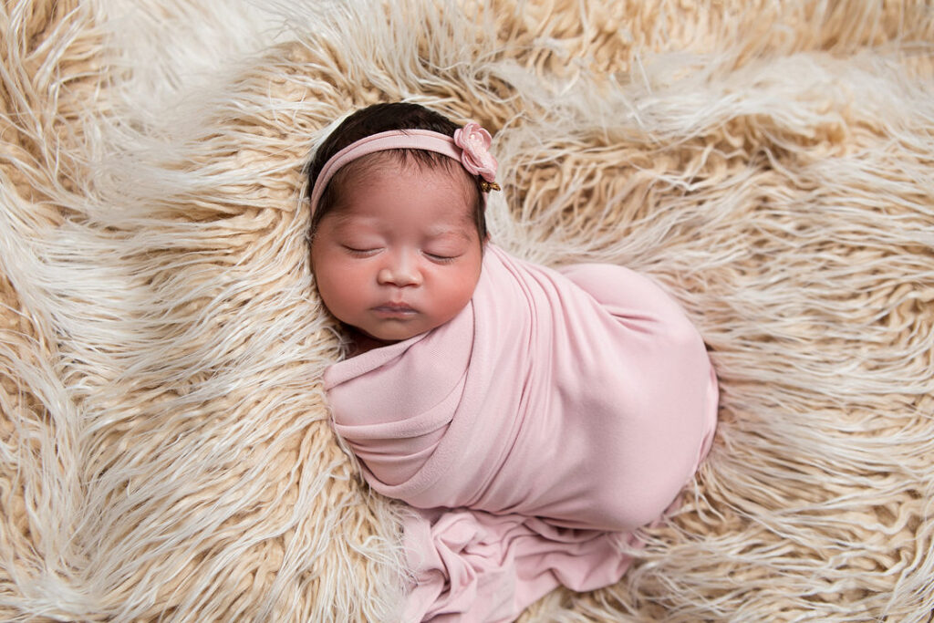 Nouveau-né emmailloté dans un tissu rose, coiffé d'un bandeau fleuri, dormant paisiblement sur une couverture duveteuse beige. Douceur et innocence.