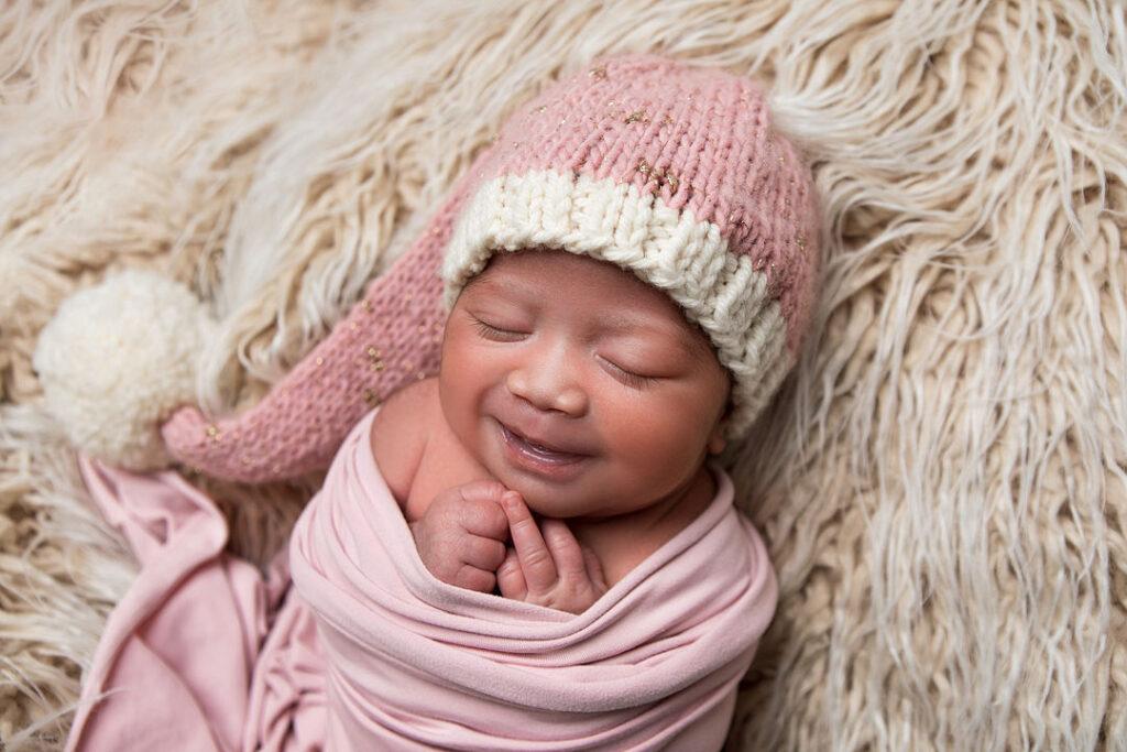 Nouveau-né emmailloté dans un tissu rose, coiffé d'un bonnet rose et blanc avec un pompon. Bébé souriant, paisible sur une couverture duveteuse.