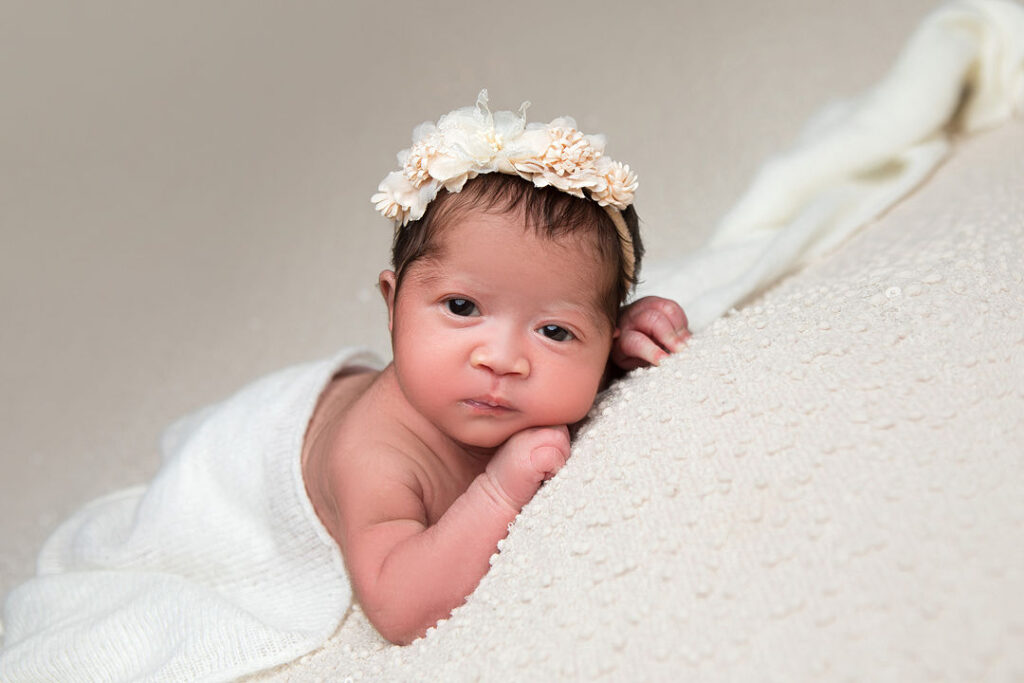 Nouveau-né paisible enveloppé dans une couverture blanche, coiffé d'une couronne florale crème. Bébé reposant sur un tissu texturé clair.