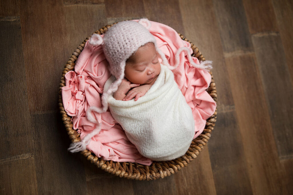 Nouveau-né paisible enveloppé dans une couverture blanche et coiffé d'un bonnet rose tricoté, dormant dans un panier sur un fond en bois chaleureux.