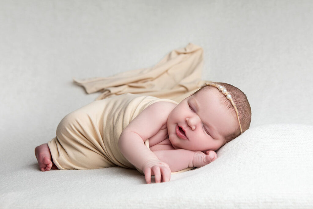 Nouveau-né dormant paisiblement, enveloppé dans un tissu beige doux, coiffé d'un fin bandeau de perles. Repose sur un coussin blanc moelleux, fond clair.