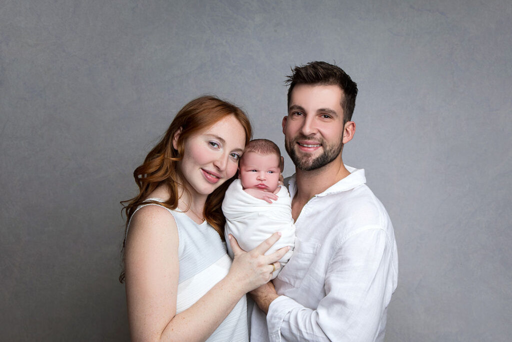 Jeune famille rayonnante : mère rousse, père brun, tenant leur nouveau-né emmailloté sur fond gris neutre.