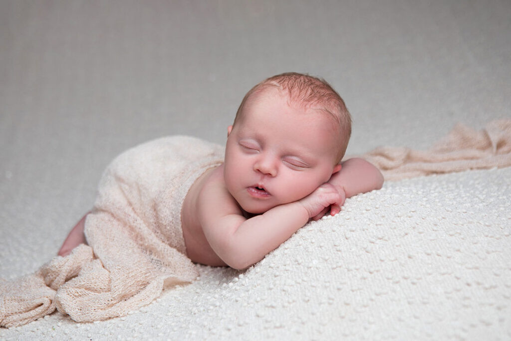 Nouveau-né paisible enveloppé dans un tissu beige, dormant sur une couverture texturée blanche. Bébé aux cheveux clairs, endormi sur le ventre, mains jointes sous le visage.