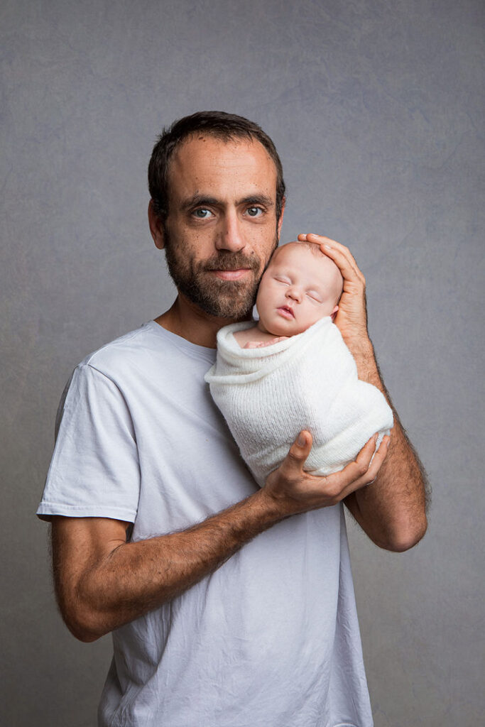 Un père tient tendrement son nouveau-né emmailloté dans un tissu blanc. L'homme, barbu, regarde la caméra avec douceur.