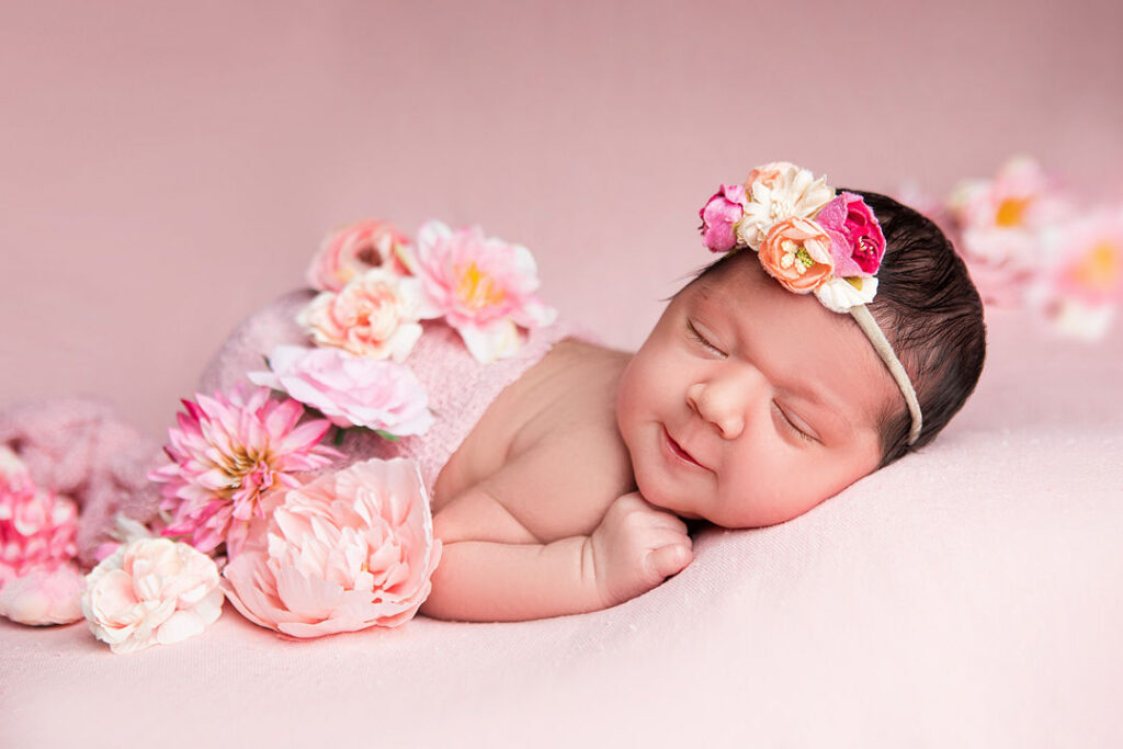 Nouveau-né dormant paisiblement sur une couverture rose, entouré de fleurs roses et coiffé d'un bandeau fleuri. Douceur et innocence capturées dans cette photo de bébé.