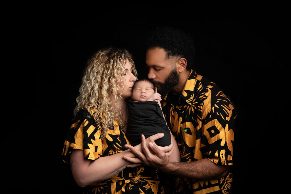 Portrait d'une famille aimante : parents en tenues assorties jaune et noir tenant leur bébé emmailloté. Le père embrasse le front du bébé, la mère regarde avec tendresse.