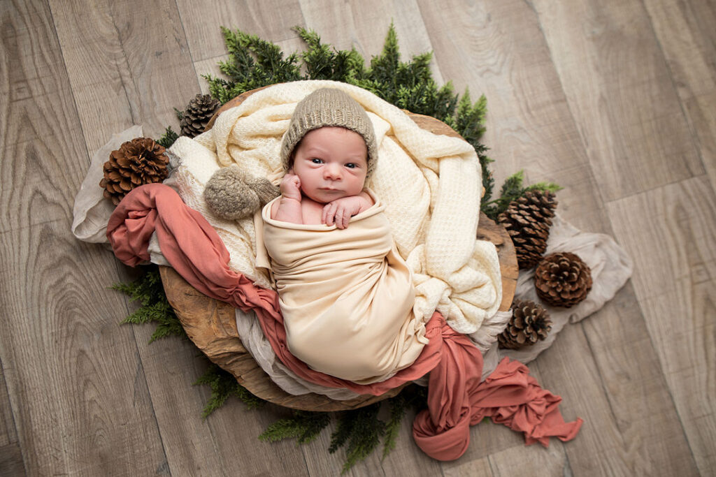 Nouveau-né emmailloté dans un panier décoré de verdure, de pommes de pin et de tissus crème et corail. Bébé avec bonnet beige. Séance photo nouveau-né.