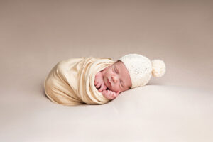 Emmaillotage ou Wrapping Nouveau-né emmailloté dans un tissu beige doux, coiffé d'un bonnet en tricot crème avec un pompon. Bébé dormant paisiblement sur un fond neutre.