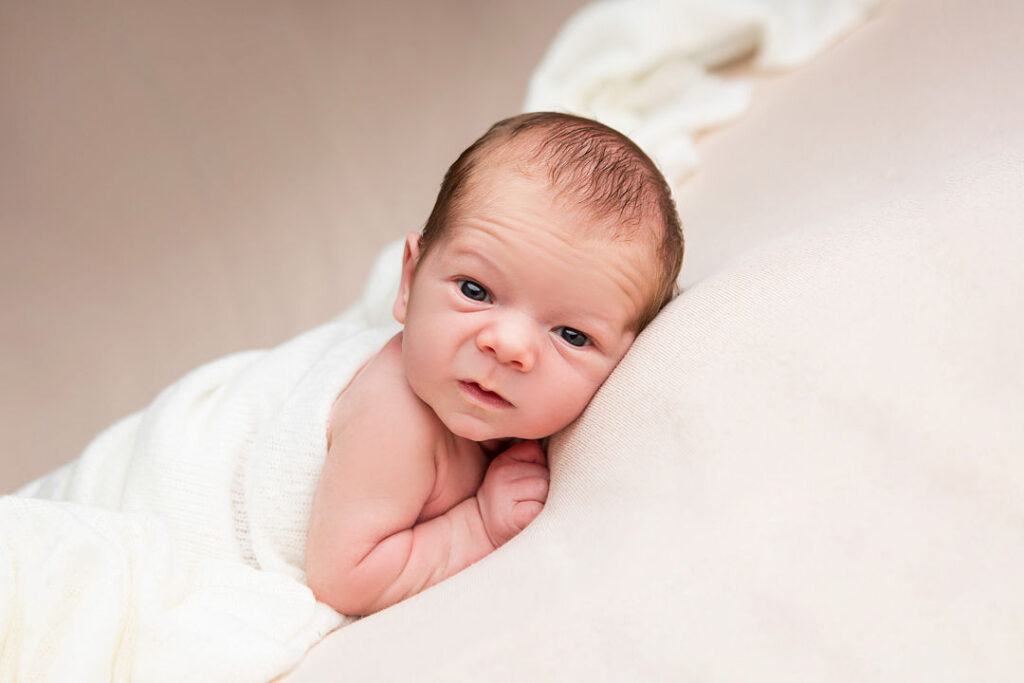 Nouveau-né enveloppé dans une couverture blanche, reposant sur un coussin beige. Le bébé a les yeux ouverts et regarde l'appareil photo.
