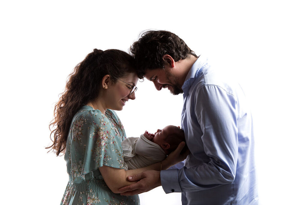 Jeune famille rayonnante: parents tenant tendrement leur nouveau-né emmailloté. La mère porte une robe fleurie turquoise. Moments précieux de parentalité.