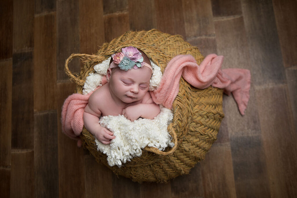 Nouveau-né dormant paisiblement dans un panier en osier, enveloppé dans une couverture blanche et un tissu rose, coiffé d'un bandeau fleuri. Séance photo bébé.