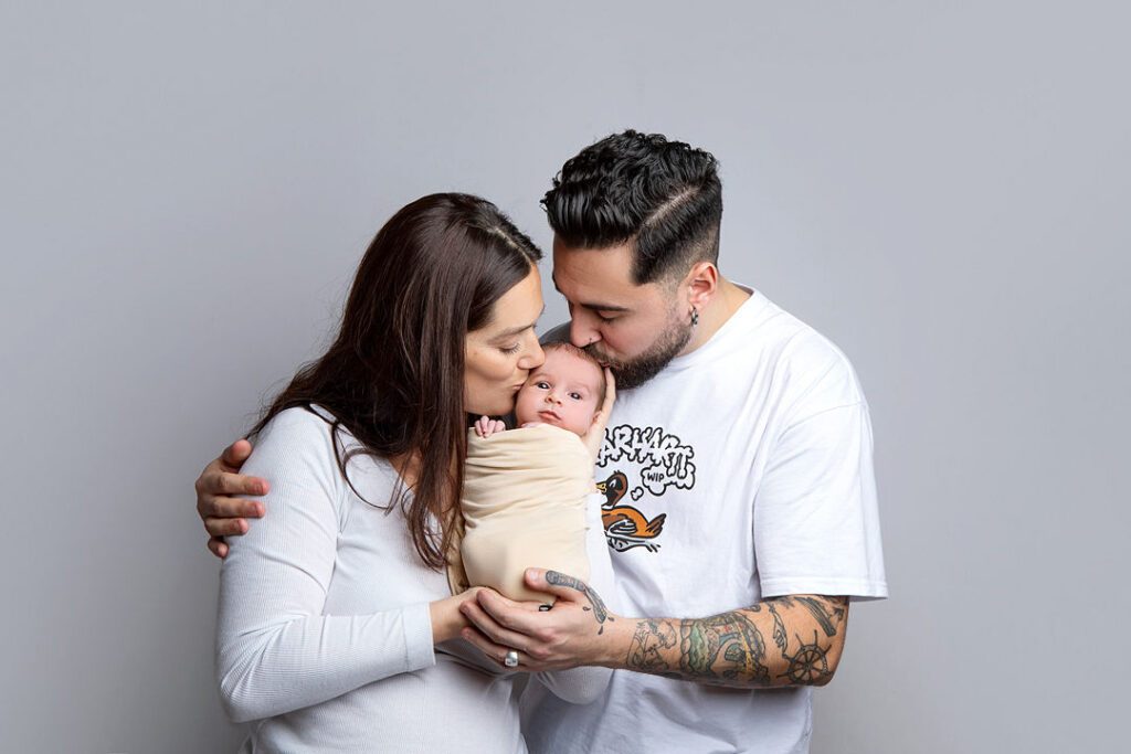 Jeune famille : parents embrassant leur nouveau-né emmailloté. Tatouages visibles, fond gris uni.