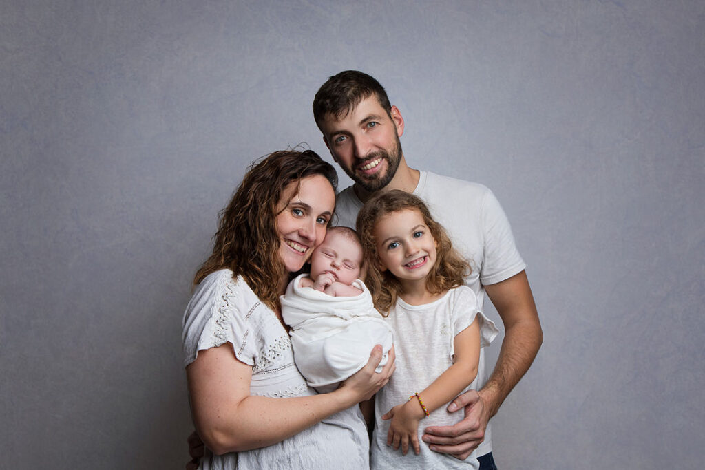 Une famille rayonnante : maman tenant un nouveau-né emmailloté, papa et fillette souriante. Fond gris doux.