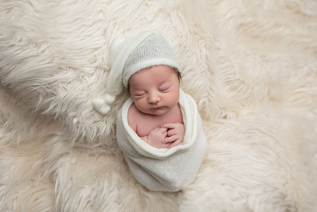 Nouveau-né emmailloté dans un lange blanc doux, coiffé d'un bonnet assorti, dormant paisiblement sur une couverture en fausse fourrure crème.