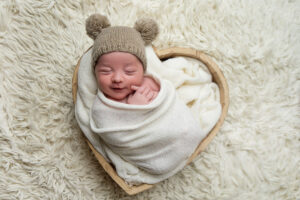 Nouveau-né paisible enveloppé dans un lange blanc, dormant dans un panier en forme de cœur sur une douce couverture duveteuse. Il porte un bonnet tricoté beige avec des pompons.