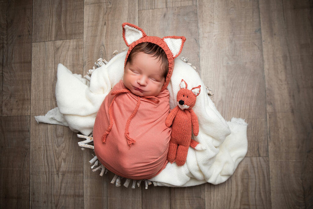 Nouveau-né emmailloté dans un tissu pêche, coiffé d'un bonnet renard, dormant paisiblement dans un panier sur un fond de bois. Un petit renard en peluche le rejoint pour une sieste câline.