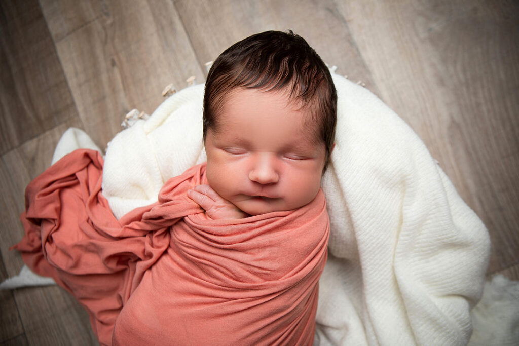 Nouveau-né emmailloté dans un tissu pêche, paisiblement endormi sur une couverture blanche et moelleuse, sur un fond de bois clair.