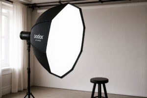 Studio photo professionnel avec un grand softbox Godox AD-S85S monté sur un trépied, prêt pour un éclairage de portrait, avec un tabouret noir à proximité et une fenêtre en arrière-plan.