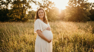 Femme enceinte souriante portant une robe longue claire dans un champ de hautes herbes au soleil couchant.